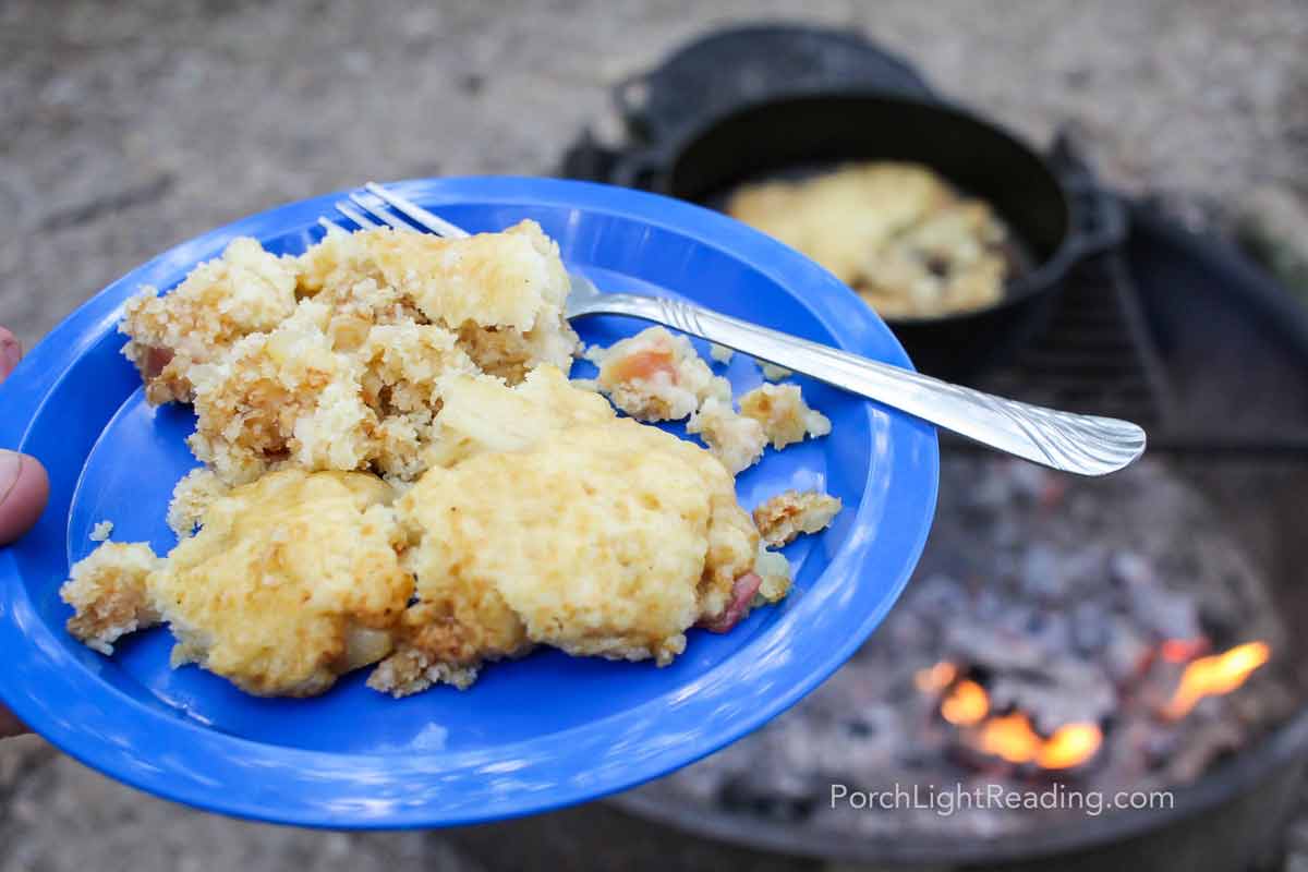 Warm apple cobbler on a blue place with a campfire and dutch oven in the background. 