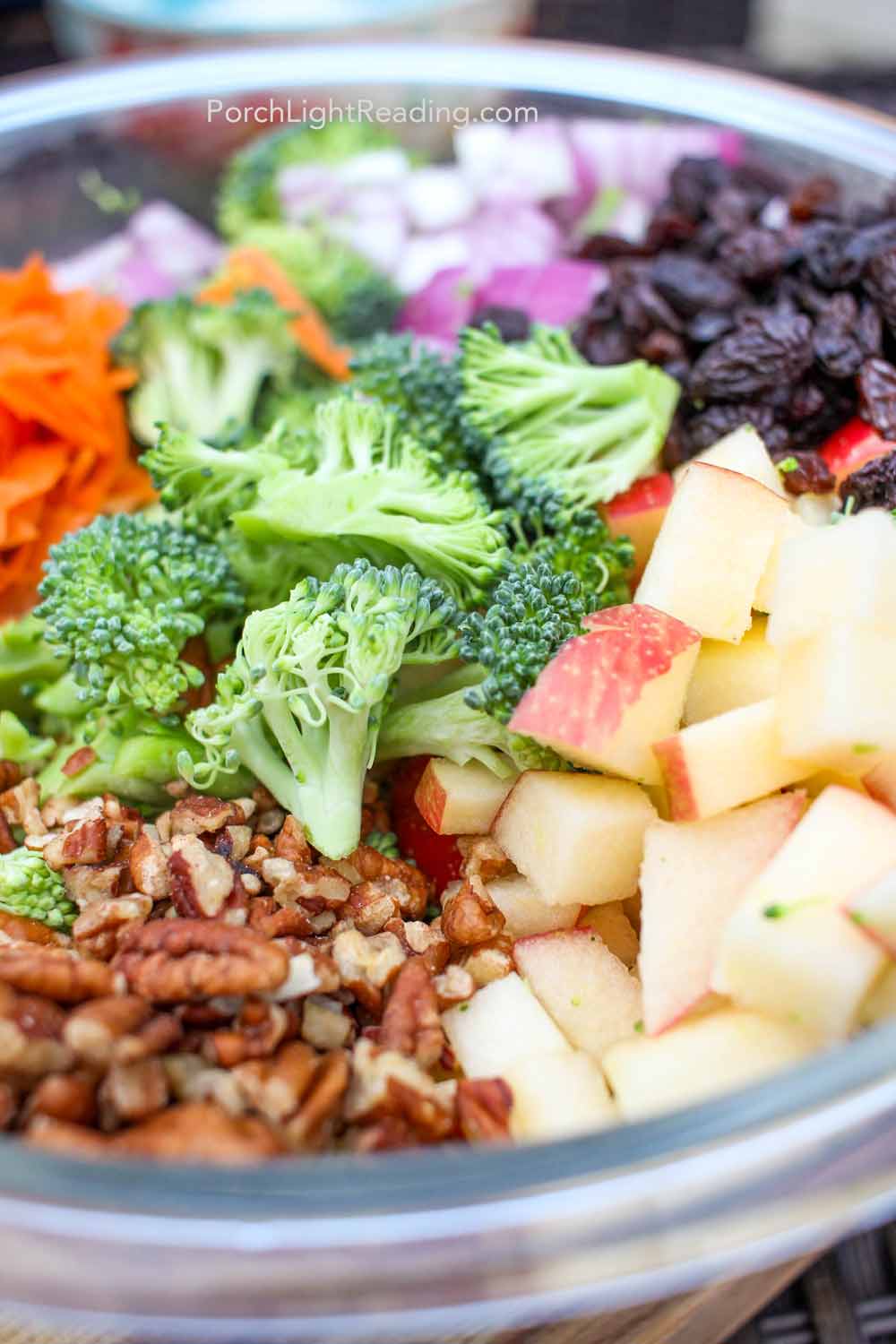 Broccoli, red onions, apple, shredded carrots, raisins and nuts in a glass bowl. 