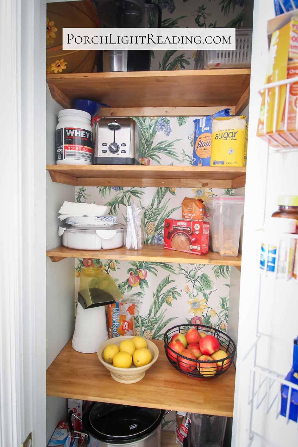 Neatly organized tiny pantry with wooden shelving. Including popcorn maker, picnic supplies, and bowls of fruit. 