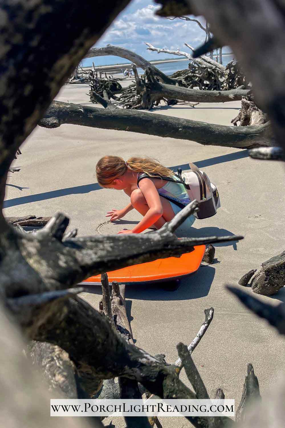 Little girl drawing in the sand surrounded by driftwood. 